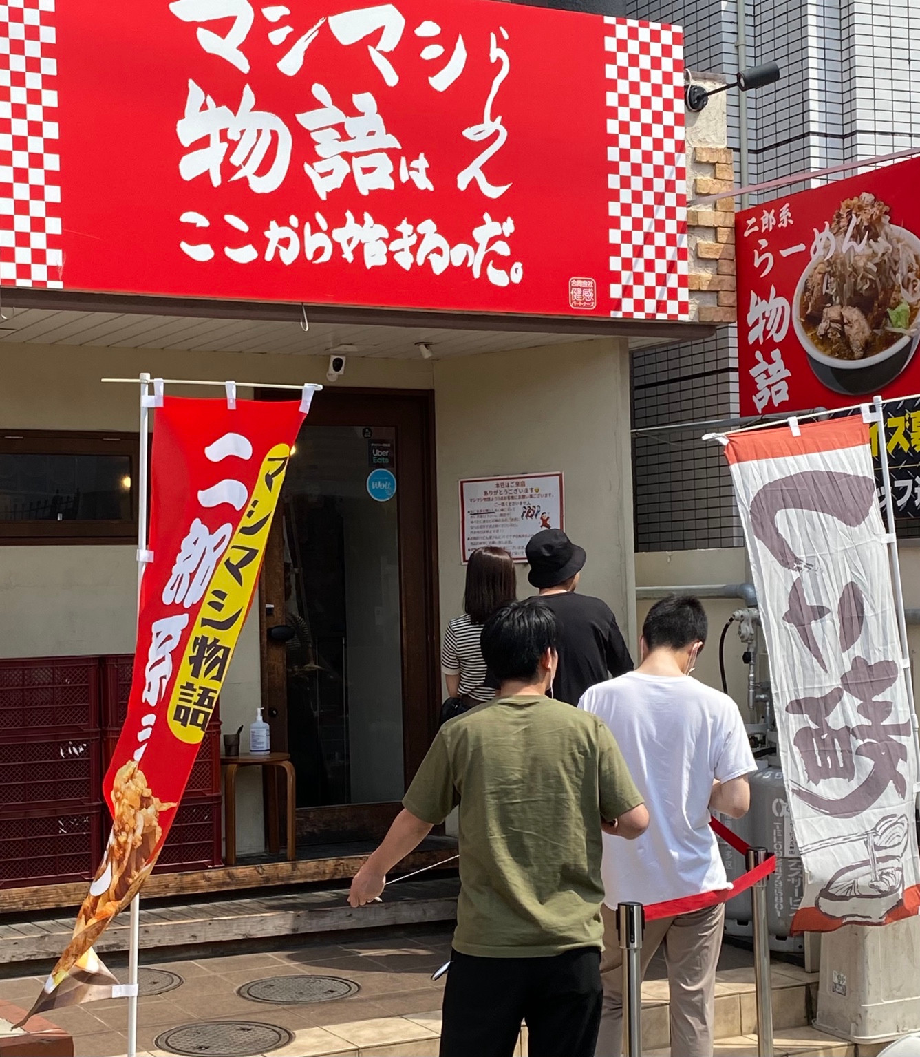 赤い看板を掲げたマシマシ物語の昼の店舗外観と行列